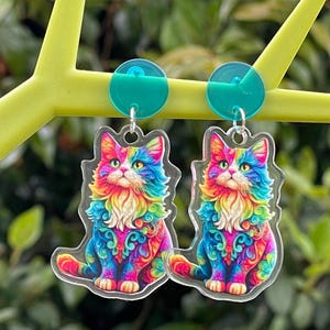 Puede incluir: Un par de pendientes con forma de gato coloridos. Cada pendiente presenta un diseño de gato sentado con vibrantes tonos arcoíris, incluyendo rojo, azul, amarillo y verde. Los pendientes están suspendidos de tachuelas circulares de color turquesa.