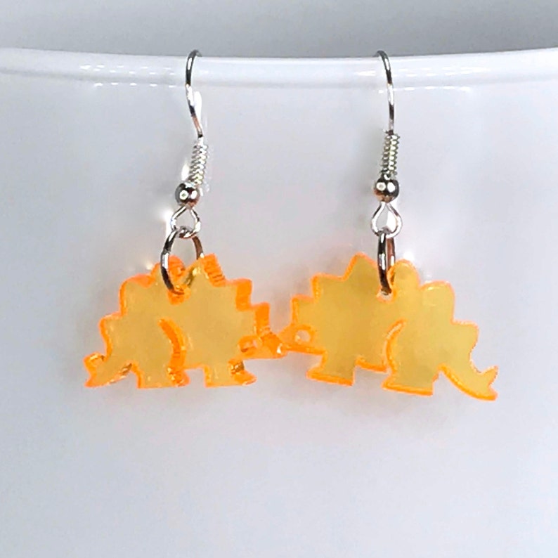 May include: A pair of yellow dinosaur earrings with silver hooks. The dinosaurs are facing each other and are made of a translucent material.