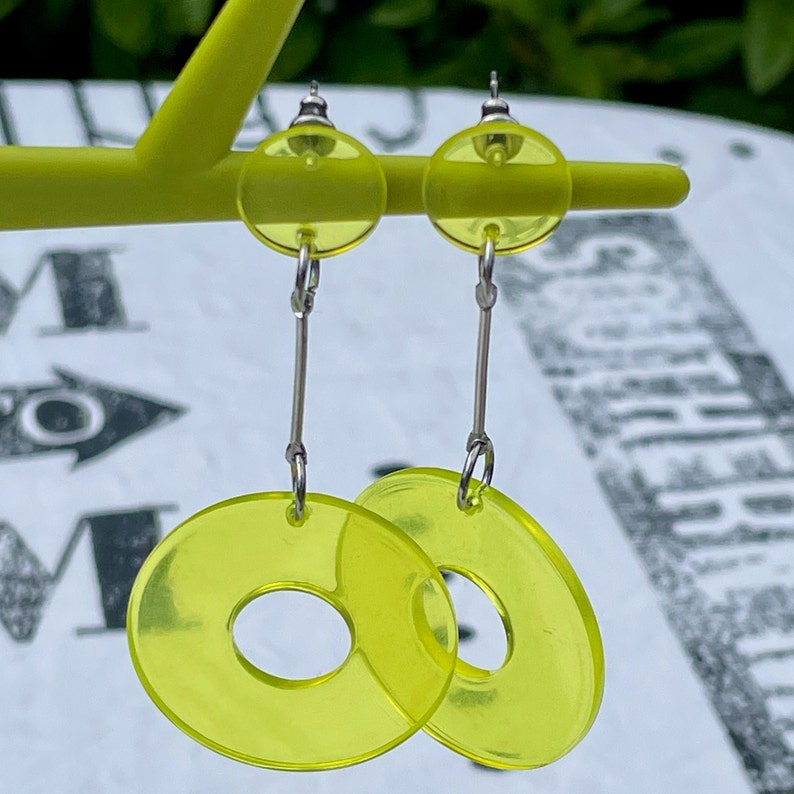 May include: A pair of yellow dangle earrings with a round, hollow center. The earrings are made of a translucent material and have a silver hook.