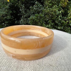 May include: A wide, round bangle bracelet in shades of amber and cream. The bracelet features a central band of cream-coloured material, with amber-coloured bands above and below. The jewellery is set against a neutral background.