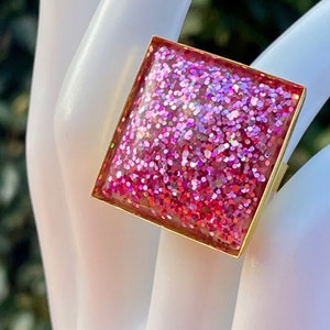 May include: A square-shaped ring with a pink glitter resin inlay and a gold metal frame.