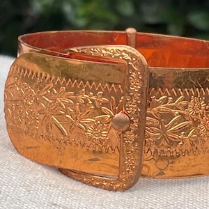 May include: A copper-colored cuff bracelet with a buckle design. The bracelet features an embossed floral pattern and a decorative buckle closure. The metal has a warm, reddish-brown hue, and the design is detailed and intricate.