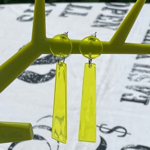 May include: A pair of yellow dangle earrings with a clear, translucent finish. The earrings are made of acrylic and have a simple, geometric design.