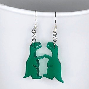 May include: A pair of green dinosaur earrings with silver hooks. The dinosaurs are facing each other and appear to be fighting.