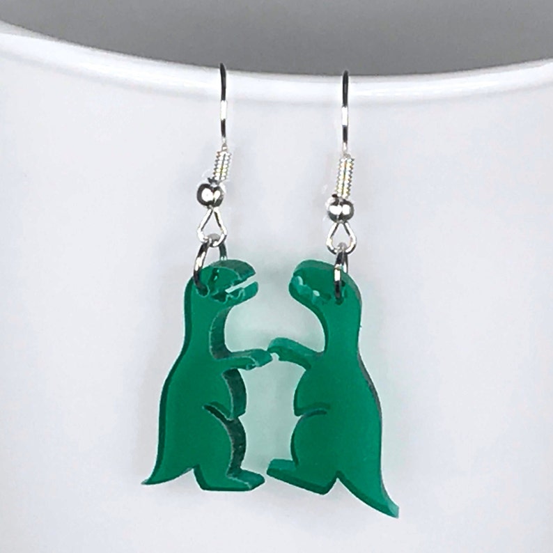 May include: A pair of green dinosaur earrings with silver hooks. The dinosaurs are facing each other and appear to be fighting.