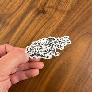 May include: A black and white line drawing sticker of a toad with a fire coming out of its mouth.
