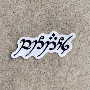 Puede incluir: Una pegatina blanca con escritura élfica negra. La pegatina tiene una forma irregular y ondulada y está colocada sobre una superficie gris texturizada. La escritura parece ser una cita o frase de una novela de fantasía.