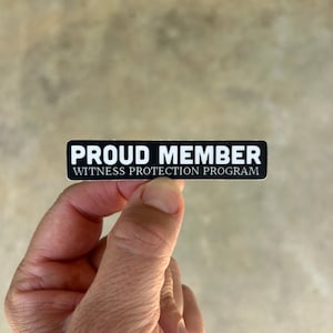 Op de afbeelding: Een rechthoekige zwarte sticker met witte tekst die PROUD MEMBER WITNESS PROTECTION PROGRAM leest in een vet, schreefloos lettertype. De sticker heeft een strak, contrastrijk ontwerp met witte letters op een effen zwarte achtergrond.
