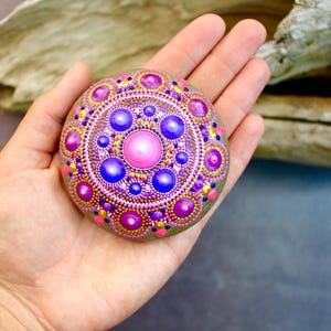 Peut inclure: Une pierre ronde et lisse ornée d'un motif mandala peint à la main dans des tons vifs de rose, violet et or. Le motif se compose de cercles concentriques de points de tailles variées, rayonnant à partir d'un point rose vif sur une base grise.