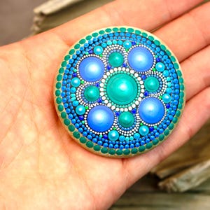 Peut inclure: Une pierre ronde et plate ornée d'un motif mandala peint à la main, composé de cercles concentriques de points dans des tons turquoise, sarcelle, bleu roi et blanc, disposés en un motif symétrique rayonnant depuis un point central vert.