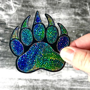 Op de afbeelding: Een zwarte berenpootafdruk sticker met een blauw, groen en regenboogkleurig glitterontwerp.