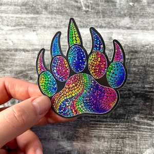 Op de afbeelding: Een regenboogkleurige, iriserende sticker met een berenpootafdruk. De poot is gemaakt van kleine, kleurrijke stippen die een mozaïek-effect creëren.