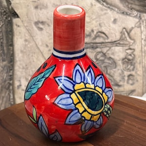 Peut inclure: Un petit vase en céramique rouge avec un intérieur blanc et un col cylindrique. Le vase est orné de motifs floraux bleus et jaunes et de feuilles vertes. Une bande bleue et blanche entoure le col. Le vase repose sur une surface en bois.