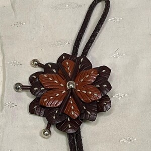 Może przedstawiać: Brązowy sk&oacute;rzany krawat bolo z kwiatowym wzorem. Kwiat ma warstwowe płatki w odcieniach brązu i beżu, z białymi przeszyciami. Srebrne metalowe akcenty znajdują się na końcach kwiatu i plecionego sznurka.