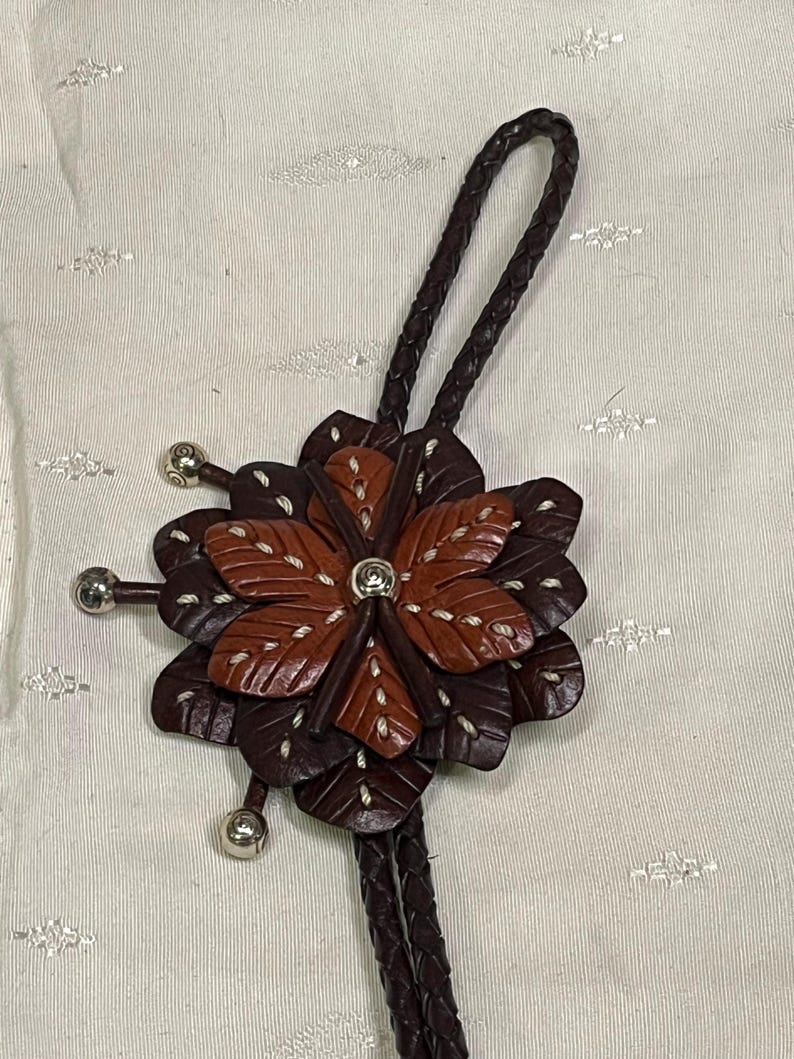 Może przedstawiać: Brązowy sk&oacute;rzany krawat bolo z kwiatowym wzorem. Kwiat ma warstwowe płatki w odcieniach brązu i beżu, z białymi przeszyciami. Srebrne metalowe akcenty znajdują się na końcach kwiatu i plecionego sznurka.