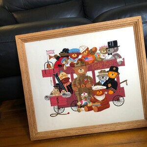 May include: A framed embroidery depicting a group of teddy bears riding a red bus. The bus is decorated with flags and the words "Teddy Bus". The bears are wearing various outfits, including military uniforms, hats, and scarves.
