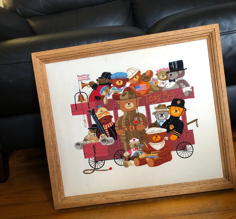 May include: A framed embroidery depicting a group of teddy bears riding a red bus. The bus is decorated with flags and the words "Teddy Bus". The bears are wearing various outfits, including military uniforms, hats, and scarves.