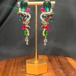 Puede incluir: Un par de pendientes largos colgantes con una montura de metal dorado incrustada con pedrería transparente. Los pendientes están decorados con cristales facetados redondos y en forma de lágrima de colores verde esmeralda, rosa intenso, azul y marrón.