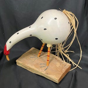 May include: A whimsical, handcrafted gourd bird sculpture. The white body is speckled with black dots, and the long neck curves down to a red beak. The bird stands on orange and yellow legs, mounted on a wooden base, with straw accents.