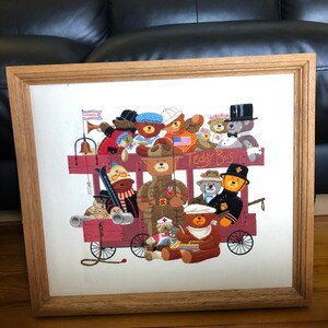 May include: A framed embroidery depicting a red wagon filled with teddy bears. The wagon is labeled "Teddy Bus" and has a bell and a flag that says "Express". The bears are dressed in various outfits, including a soldier, a sailor, a doctor, and a chef.