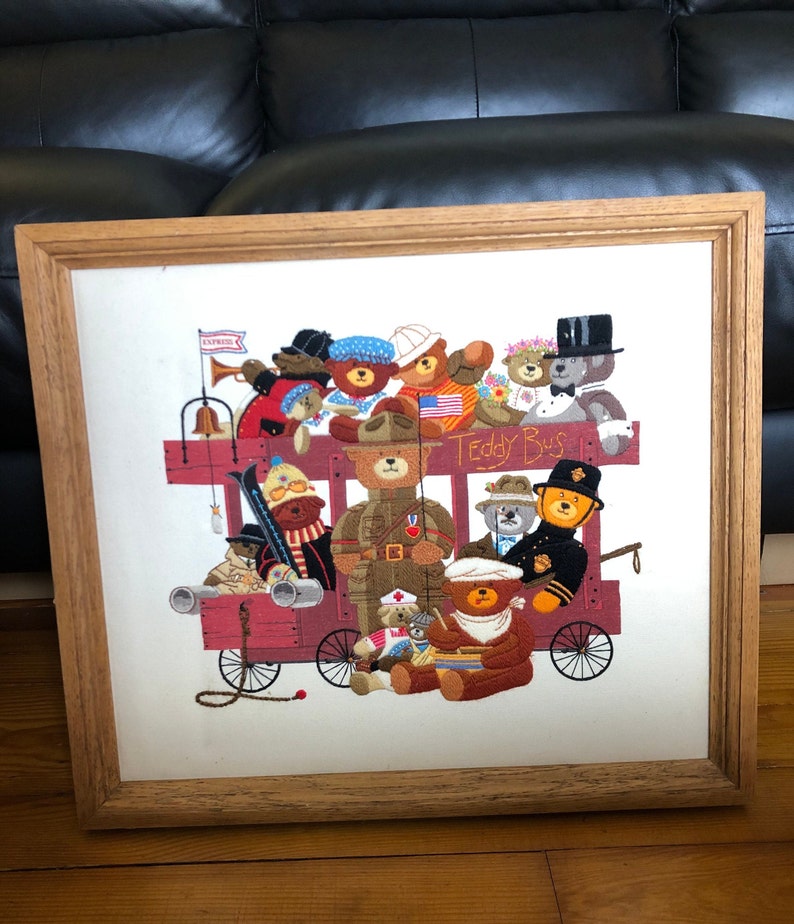 May include: A framed embroidery depicting a red wagon filled with teddy bears. The wagon is labeled "Teddy Bus" and has a bell and a flag that says "Express". The bears are dressed in various outfits, including a soldier, a sailor, a doctor, and a chef.