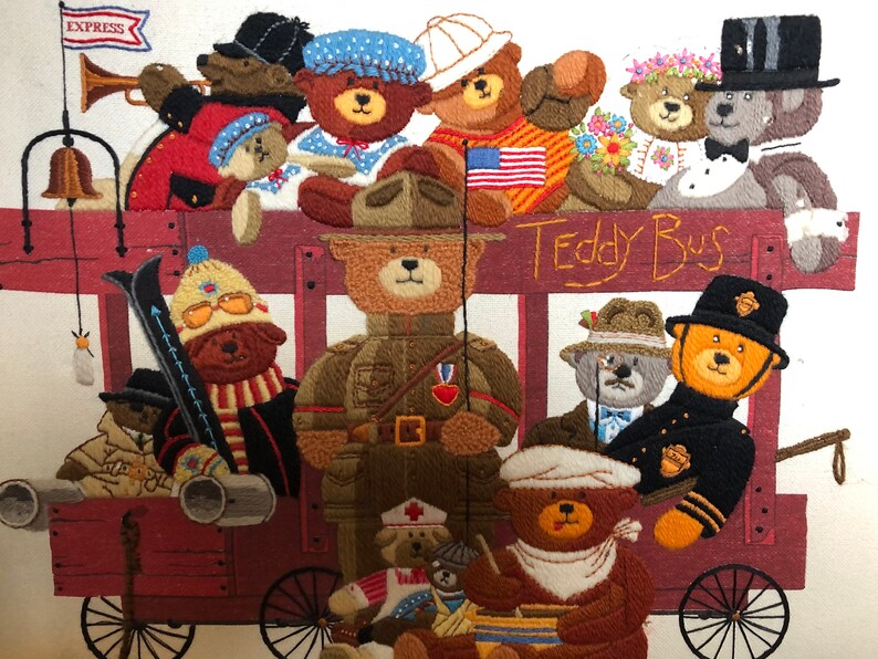 May include: A colorful embroidered picture of a teddy bear bus with various teddy bears dressed in different outfits. The bus is red with a wooden frame and wheels. The teddy bears are wearing hats, scarves, and other accessories. The text "Teddy Bus" is embroidered on the side of the bus.