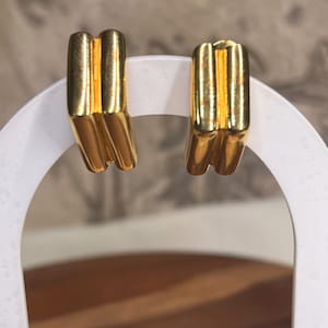 May include: A pair of gold-colored earrings. Each earring has a rectangular shape with three vertical lines on the front. The earrings are displayed on a white stand against a blurred background.
