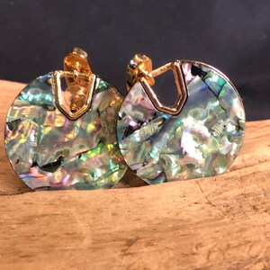 May include: A pair of circular earrings featuring iridescent abalone shell inlays with a shimmering mix of green, blue, and purple tones. Each earring is accented with a polished gold-tone metal frame that includes a V-shaped cutout at the top.