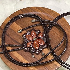 Może przedstawiać: Brązowy krawat bolo z rzeźbioną drewnianą zawieszką w kształcie kwiatu. Zawieszka posiada centralny kryształ i otoczona jest ciemnobrązowymi i czerwonobrązowymi płatkami. Pleciony sk&oacute;rzany sznurek jest ciemnobrązowy.