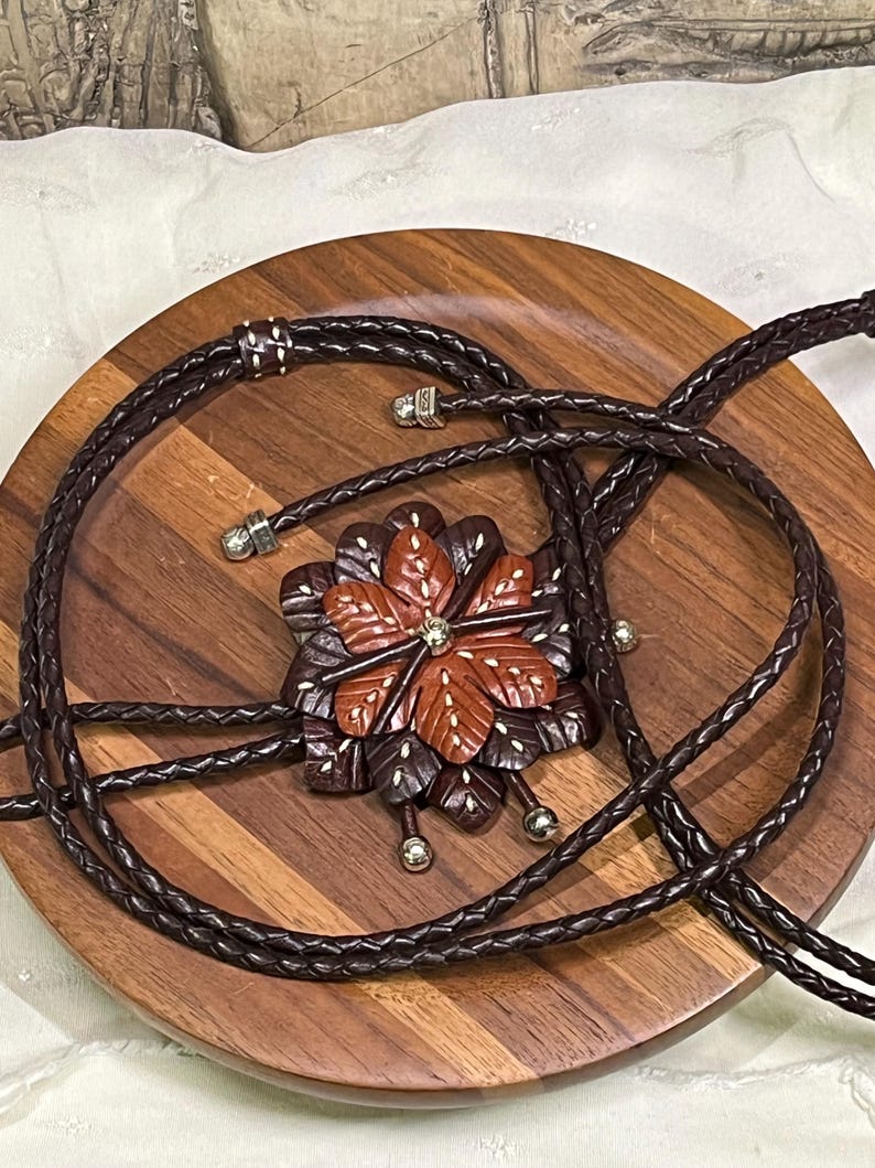 Może przedstawiać: Brązowy krawat bolo z rzeźbioną drewnianą zawieszką w kształcie kwiatu. Zawieszka posiada centralny kryształ i otoczona jest ciemnobrązowymi i czerwonobrązowymi płatkami. Pleciony sk&oacute;rzany sznurek jest ciemnobrązowy.