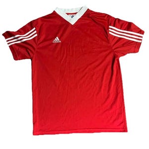 K&ouml;nnte beinhalten: Rotes Sporttrikot mit wei&szlig;em V-Ausschnitt und wei&szlig;en Streifen auf den &Auml;rmeln. Das Adidas-Logo befindet sich auf der Brust. Dieses Vintage-Fu&szlig;balltrikot ist ein klassischer Sportartikel.