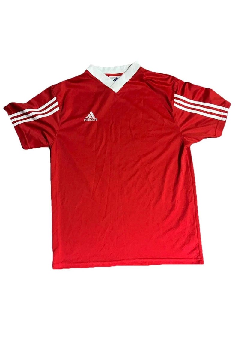 K&ouml;nnte beinhalten: Rotes Sporttrikot mit wei&szlig;em V-Ausschnitt und wei&szlig;en Streifen auf den &Auml;rmeln. Das Adidas-Logo befindet sich auf der Brust. Dieses Vintage-Fu&szlig;balltrikot ist ein klassischer Sportartikel.