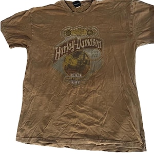 May include: Brown Harley-Davidson t-shirt with a distressed appearance. The front displays a Harley-Davidson logo with the text "Since 1903" and "Motor Harley-Davidson Cycles". The shirt has a crew neck and short sleeves.