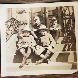 May include: Black and white vintage photograph of five children sitting on a set of stairs. The children are wearing hats and coats. The photo is taken in front of a building with a wooden door.