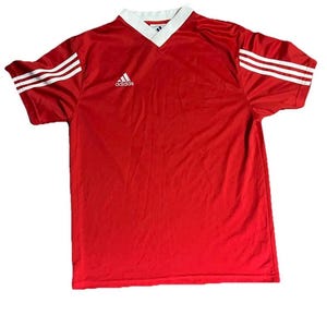 K&ouml;nnte beinhalten: Rotes Adidas Fu&szlig;balltrikot mit wei&szlig;em V-Ausschnitt und wei&szlig;en Streifen auf den &Auml;rmeln. Das Adidas-Logo befindet sich auf der Brust. Das Trikot hat eine einfarbige rote Farbe.