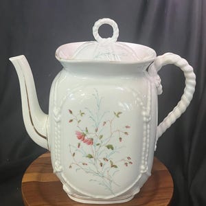 Puede incluir: Una tetera de porcelana blanca con un delicado diseño floral de flores rosas y tallos verdes. La tetera tiene un cuerpo cuadrado con detalles en relieve que imitan una cuerda en los bordes, un asa trenzada y una tapa con un remate circular.