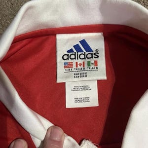 K&ouml;nnte beinhalten: Nahaufnahme eines roten Adidas-Trikots mit wei&szlig;em Kragen. Das Adidas-Logo ist auf einem Etikett zu sehen, zusammen mit Gr&ouml;&szlig;en- und Materialangaben. Das Etikett enth&auml;lt auch den Text "MADE IN MEXICO".