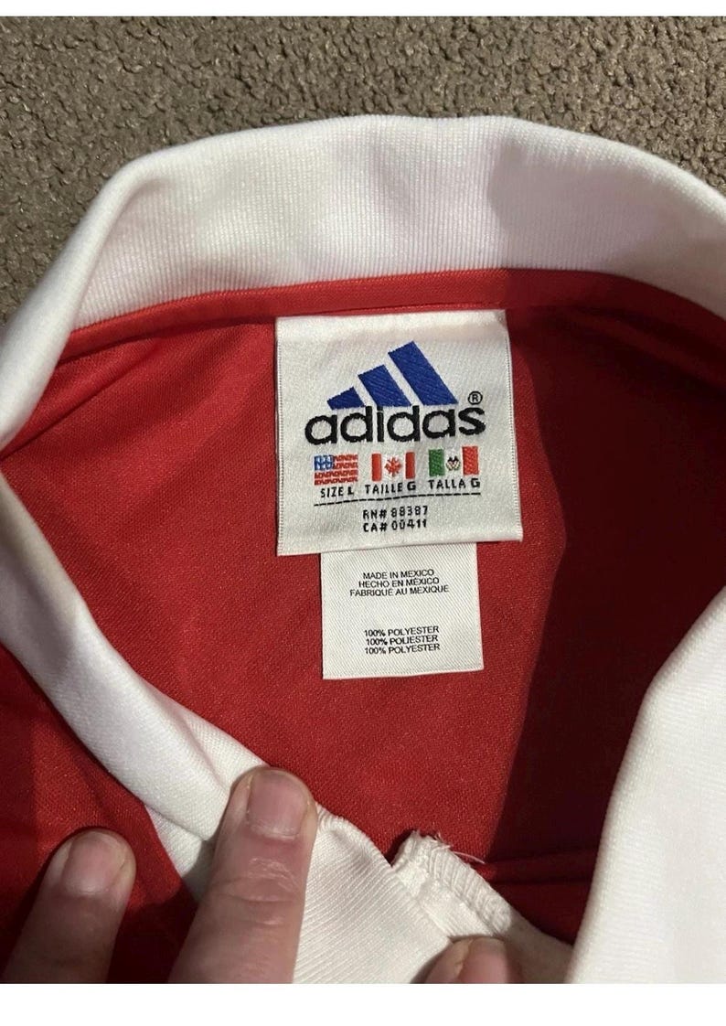 K&ouml;nnte beinhalten: Nahaufnahme eines roten Adidas-Trikots mit wei&szlig;em Kragen. Das Adidas-Logo ist auf einem Etikett zu sehen, zusammen mit Gr&ouml;&szlig;en- und Materialangaben. Das Etikett enth&auml;lt auch den Text "MADE IN MEXICO".
