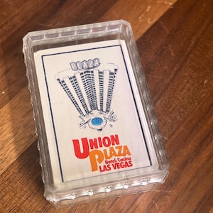 Könnte beinhalten: Ein Kartenspiel in einer durchsichtigen, strukturierten Kunststoffhülle. Die oberste Karte zeigt eine schwarz-weiße Illustration des Plaza-Hotelgebäudes mit dem Schriftzug "PLAZA" darüber und "UNION PLAZA Hotel/Casino LAS VEGAS" darunter.