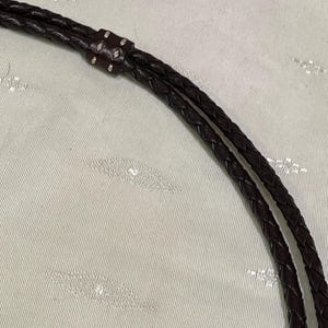 Może przedstawiać: Zbliżenie na ciemnobrązowy pleciony sk&oacute;rzany pasek. Pasek jest połączony małym, ciemniejszym brązowym kawałkiem sk&oacute;ry ze srebrnymi nitami. Sk&oacute;ra wydaje się gładka i dobrze wykonana.