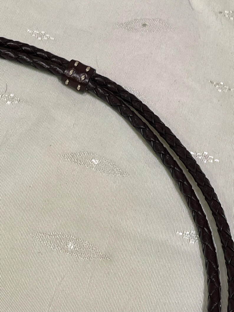 Może przedstawiać: Zbliżenie na ciemnobrązowy pleciony sk&oacute;rzany pasek. Pasek jest połączony małym, ciemniejszym brązowym kawałkiem sk&oacute;ry ze srebrnymi nitami. Sk&oacute;ra wydaje się gładka i dobrze wykonana.