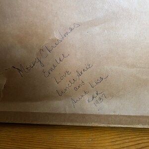 May include: A handwritten note on a brown paper background. The note says "Merry Christmas Emma Lee Love Uncle Arnie and his Aunt Kay 1987".