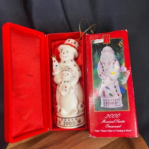 Peut inclure: Ornement de bonhomme de neige en céramique blanche avec un chapeau rouge et blanc, tenant un petit sapin de Noël. L'ornement est dans une boîte rouge avec un ornement de Père Noël assorti. Le texte sur la boîte indique "2000 Musical Santa Ornament".