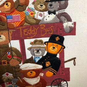 May include: A colorful embroidered picture of teddy bears riding a red wagon with the words "Teddy Bus" on the side. The bears are dressed in different outfits, including a bride and groom, a soldier, a police officer, and a doctor.