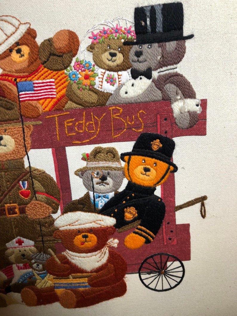 May include: A colorful embroidered picture of teddy bears riding a red wagon with the words "Teddy Bus" on the side. The bears are dressed in different outfits, including a bride and groom, a soldier, a police officer, and a doctor.