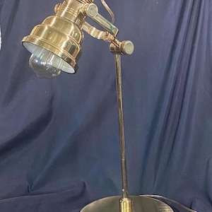 May include: A brass-toned desk lamp with an adjustable head and a round base. The lamp features a clear bulb and a flexible arm, allowing for directional lighting. The design is vintage-inspired.