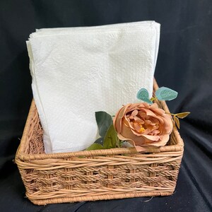 May include: A woven wicker basket containing a stack of white paper napkins and a single artificial rose with brown petals.