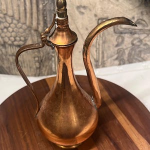 May include: A metallic copper-toned ewer featuring a slender neck, a curved spout, and a hinged lid with a pointed finial. The vessel has a rounded base and a curved handle attached to the neck, displaying a polished finish with visible patina.