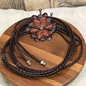 Może przedstawiać: Brązowy sk&oacute;rzany krawat bolo z rzeźbioną zawieszką w kształcie kwiatu. Zawieszka ma centralny kwiat w kolorze czerwono-brązowym otoczony ciemnobrązowymi płatkami. Pleciony sk&oacute;rzany sznurek ma srebrne końc&oacute;wki i akcenty.