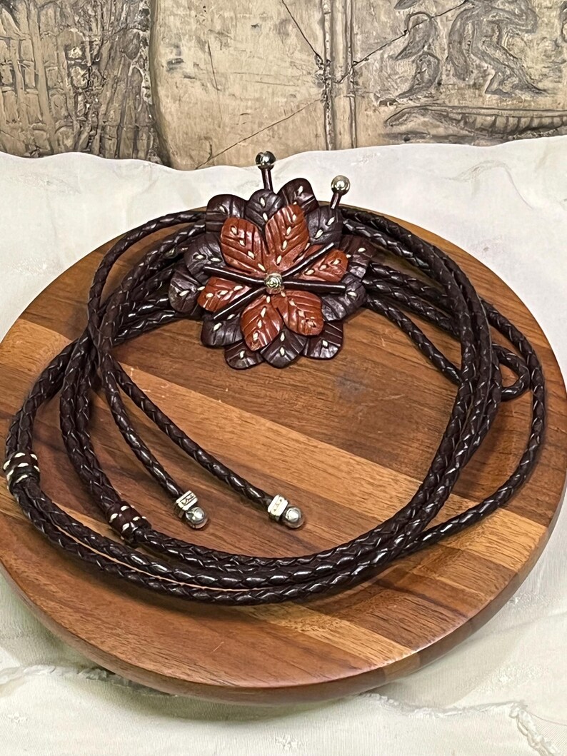 Może przedstawiać: Brązowy sk&oacute;rzany krawat bolo z rzeźbioną zawieszką w kształcie kwiatu. Zawieszka ma centralny kwiat w kolorze czerwono-brązowym otoczony ciemnobrązowymi płatkami. Pleciony sk&oacute;rzany sznurek ma srebrne końc&oacute;wki i akcenty.